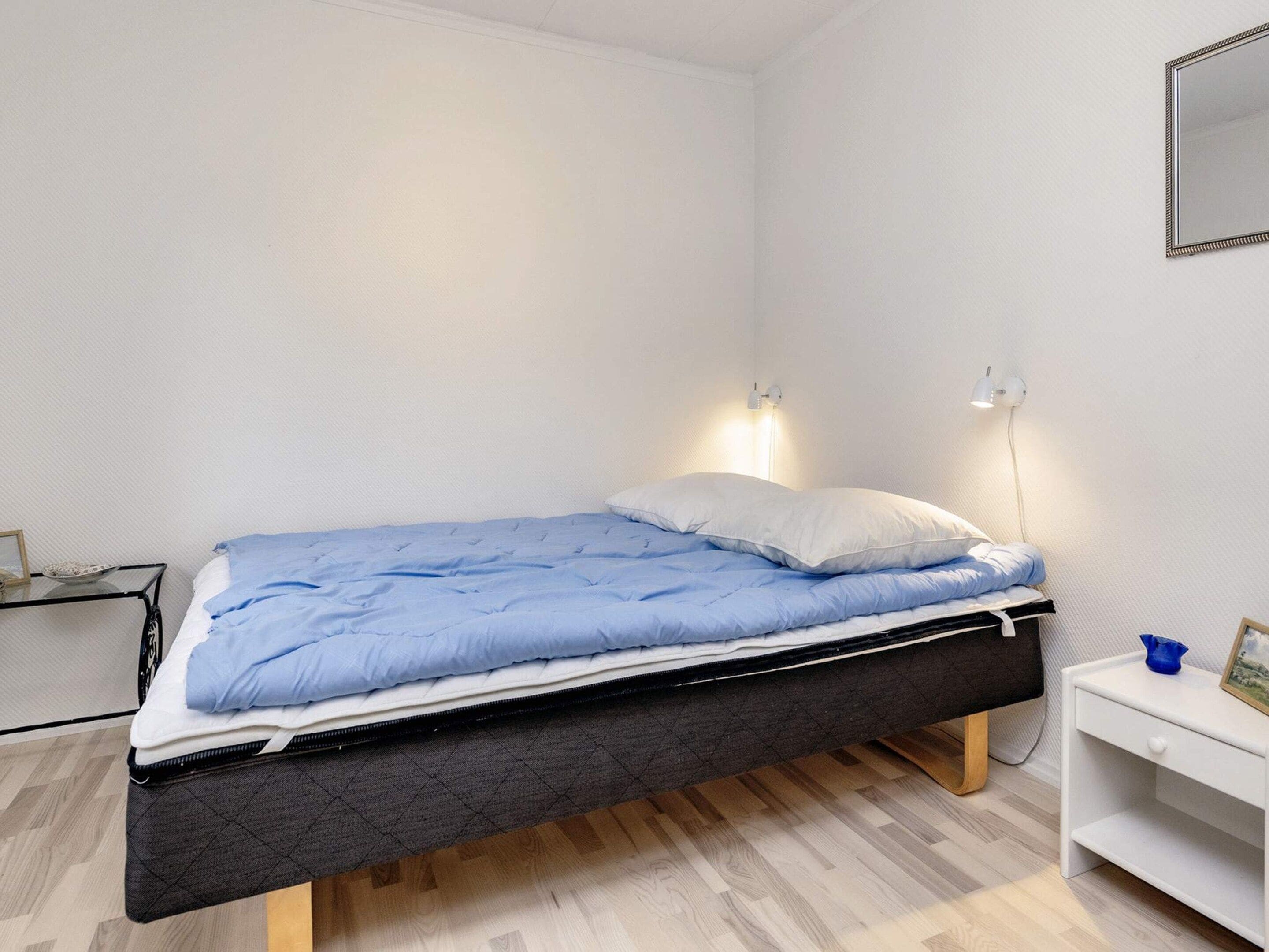 Un lit simple avec un matelas bleu et des oreillers blancs, une table de chevet avec une lampe, et une petite étagère avec un cadre photo et un objet bleu.