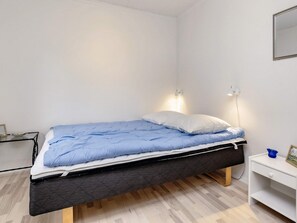 A single bed with a blue mattress and white pillows, a bedside table with a lamp, and a small shelf with a picture frame and a blue object.