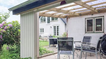 Un patio cubierto con mesa y sillas, una parrilla y un jardín con plantas y flores.