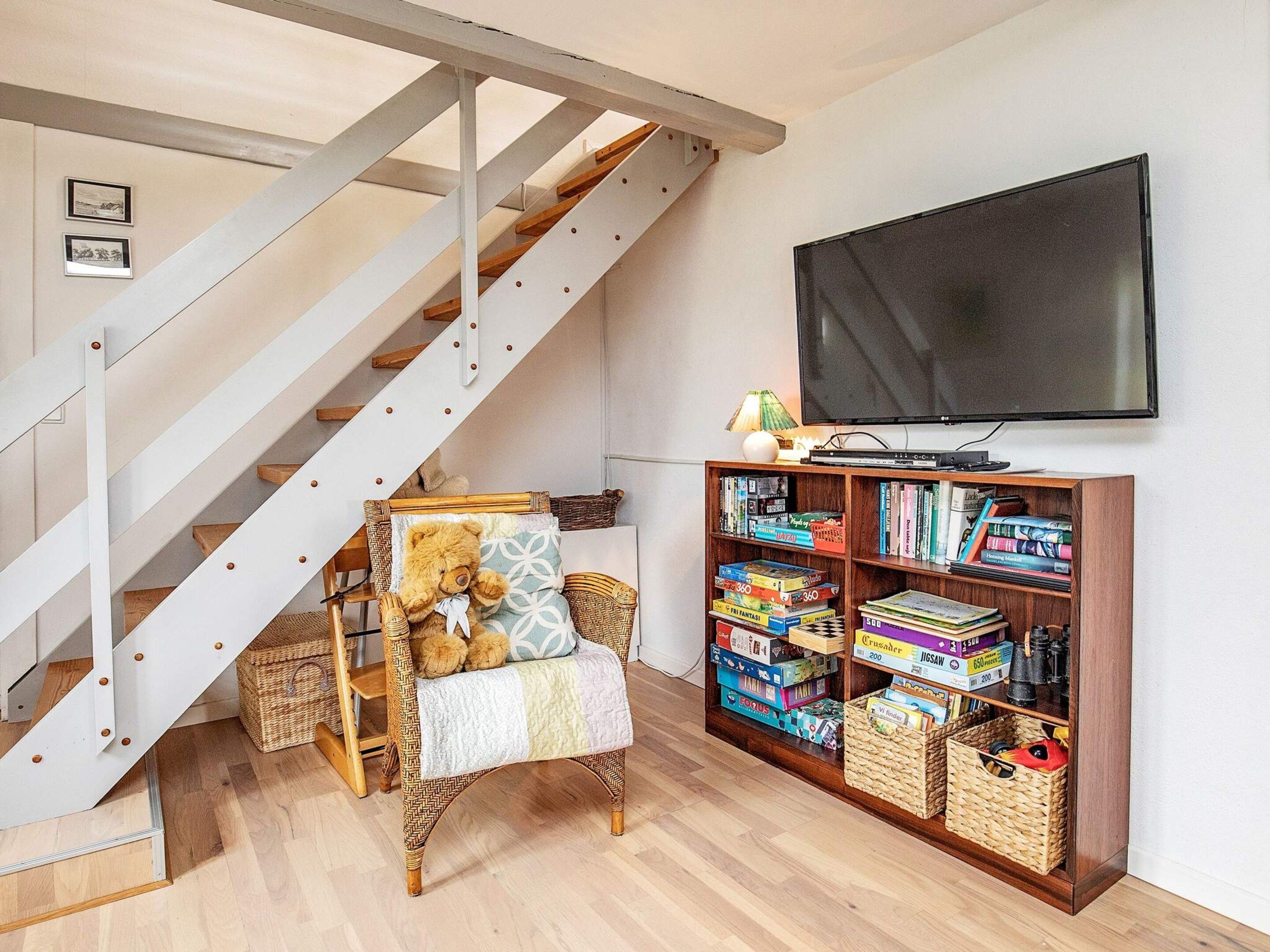 Una escalera con barandal blanco, una silla de mimbre con un osito de peluche, una estantería llena de libros y juegos, y un televisor en la pared.