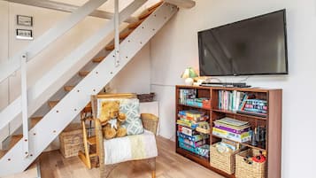 Una escalera con barandal blanco, una silla de mimbre con un osito de peluche, una estantería llena de libros y juegos, y un televisor en la pared.