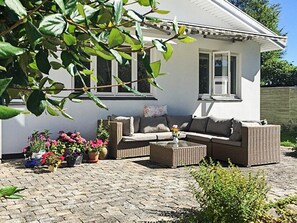 A house with a white exterior, a brown wicker sofa, and a coffee table with a vase. There are potted plants and flowers around the seating area. - Holiday Home in DronningmÃ¸lle (Dronningmolle)