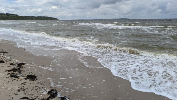 Une plage avec des vagues, un ciel nuageux et une terre lointaine à l’horizon.