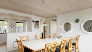 Un coin repas avec une table blanche et des chaises en bois, une cuisine en arrière-plan et un miroir rond accroché au mur.