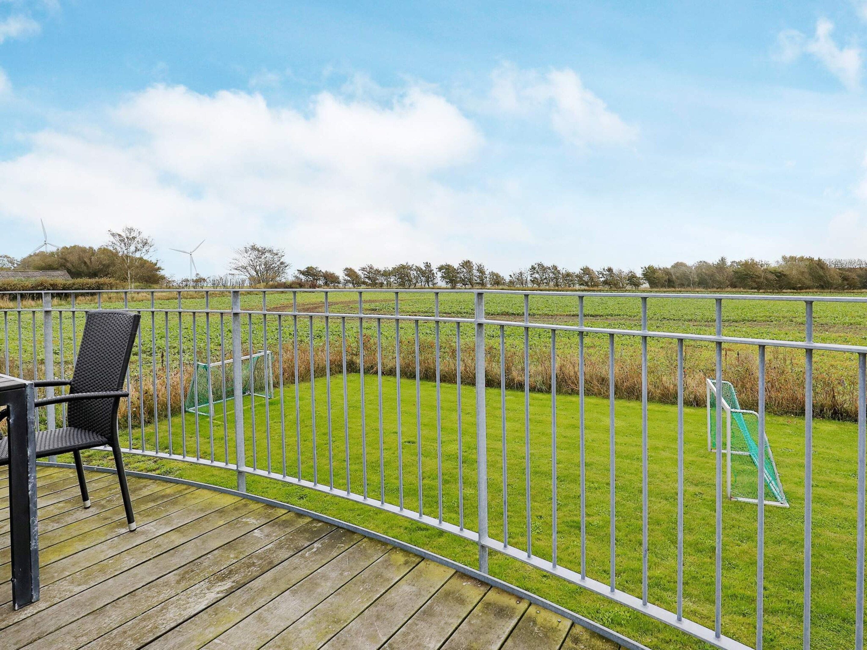 A wooden deck with a metal railing, a black chair, and a view of a green field with wind turbines in the distance.