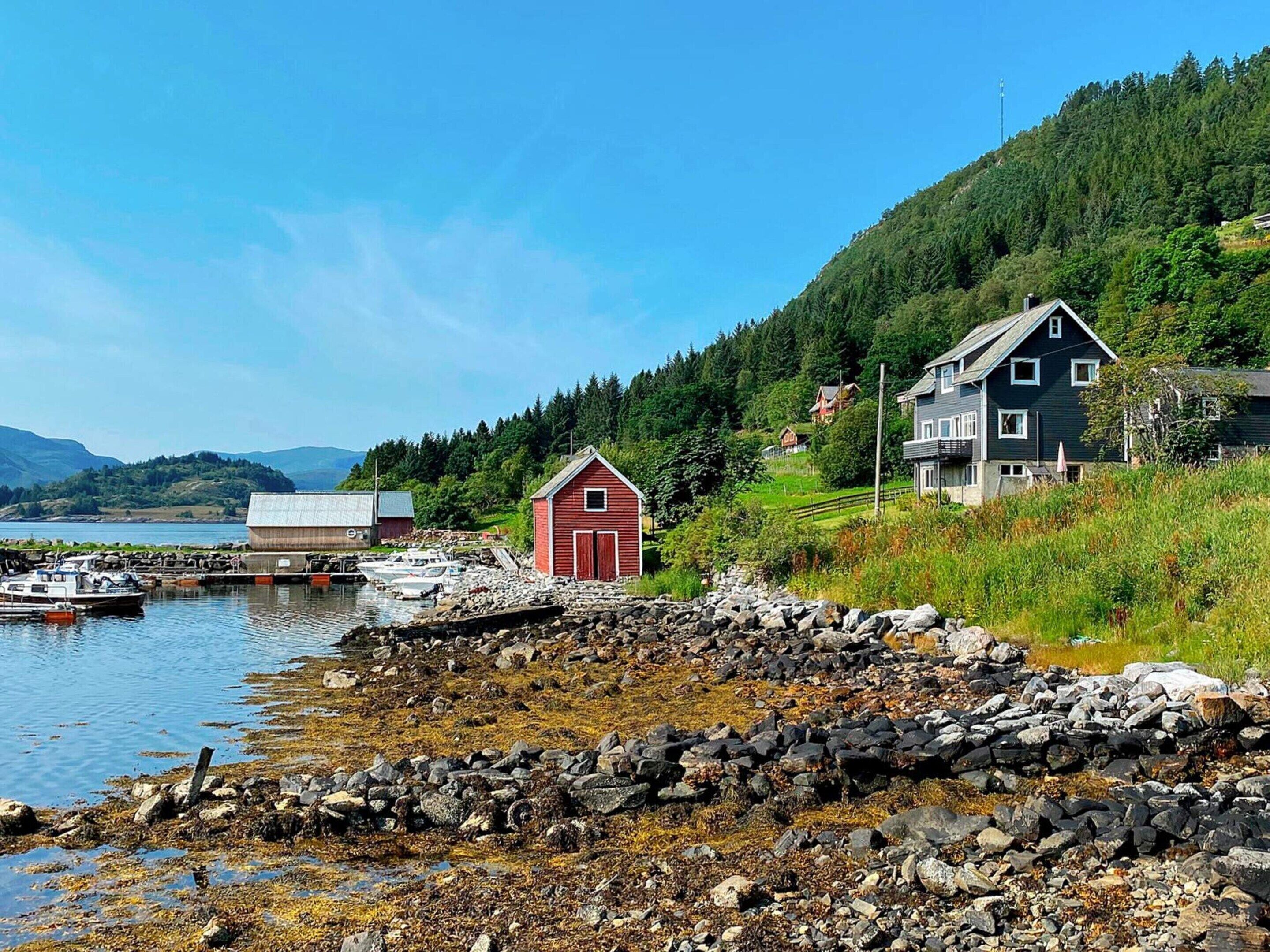 En kustvy med en röd ladugård, ett hus och båtar förtöjda vid en brygga.