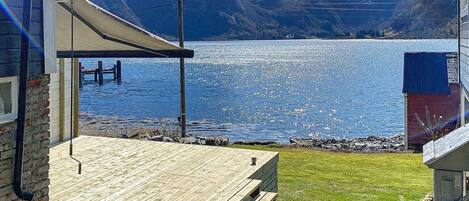 Um deck de madeira com mesa e cadeiras, com vista para um lago e montanhas ao fundo.