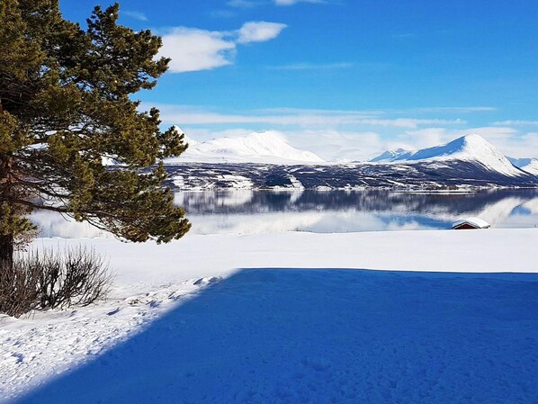 Schneebedeckte Landschaft mit einem klaren See, Bergen im Hintergrund und einer Hütte.