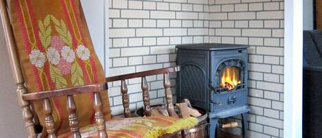 A corner room with a brick wall, a wooden rocking chair, a small stove, and a fireplace.