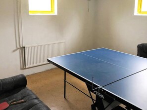 Ein Zimmer mit Tischtennisplatte, Heizkörper, einer Couch und zwei Fenstern mit gelben Rahmen.