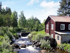 A rustic wooden house with a stone foundation, a small bridge over a flowing stream, and lush greenery in the background. - 10 Person Holiday Home in Karbole (Kårböle)