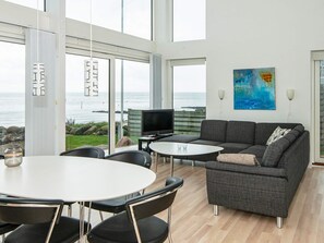 A modern living room with a large window overlooking the sea, a grey sofa, a small table, and a TV.
