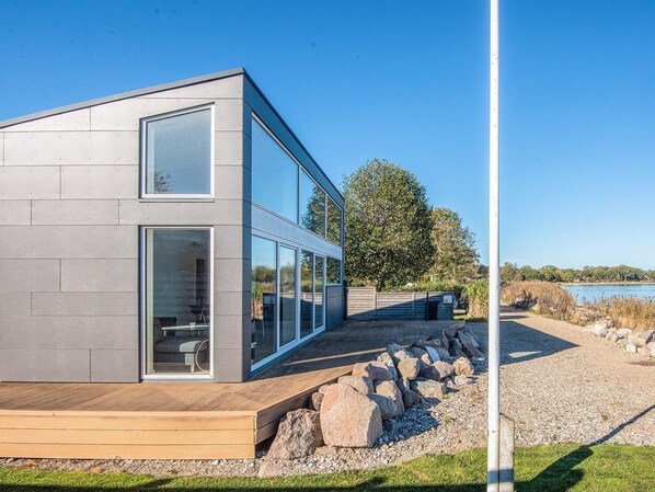 A modern building with large glass windows, a wooden deck, and a view of a lake.
