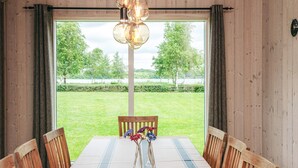 Un comedor con una mesa dispuesta para cuatro, sillas de madera, un jarrón con flores y vistas a un paisaje cubierto de hierba a través de grandes ventanales.