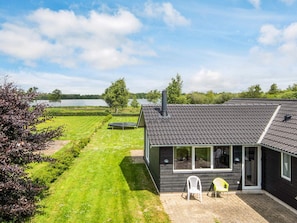 A modern house with a large window, a patio with two chairs, and a view of a lake. - Holiday Home in Ansager (Ansager)