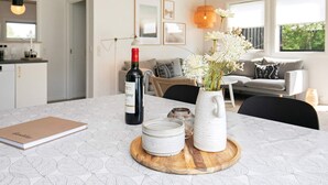 Uma sala de estar moderna com uma mesa de jantar, uma garrafa de vinho, um vaso com flores e uma cozinha ao fundo.