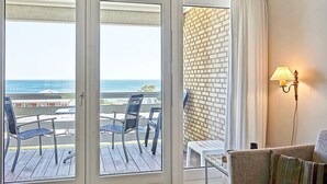 Una stanza con una porta vetrata scorrevole che dà su un patio esterno con vista sull'oceano. All'esterno c'è un tavolo e delle sedie, e all'interno una lampada a parete.