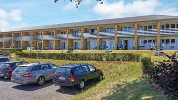 Un hotel de varios pisos con techo plano y una fila de balcones. Varios coches estacionados en la parte delantera, y el hotel cuenta con un césped y setos bien cuidados.