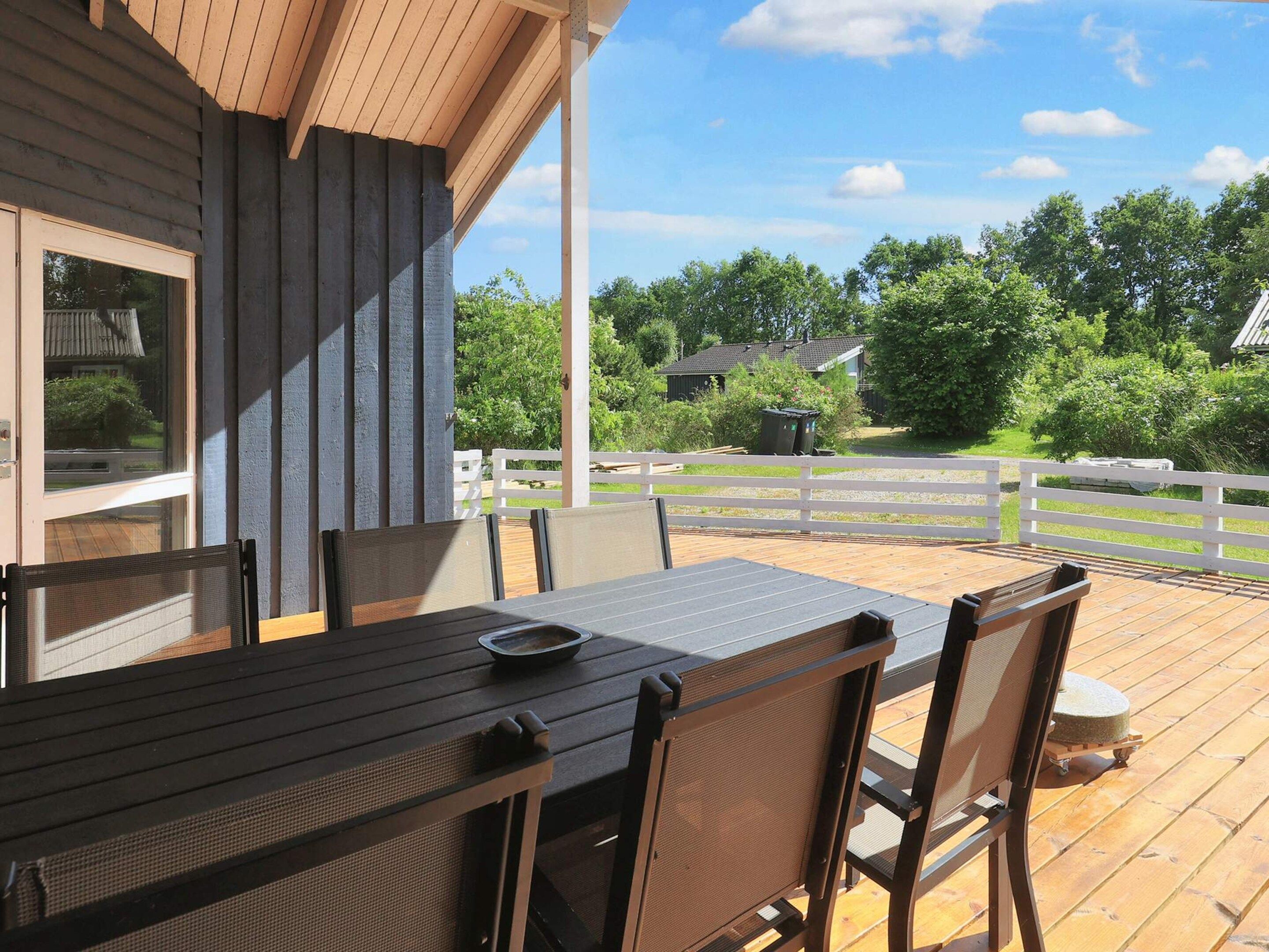 An outdoor deck with a wooden table and chairs, a white fence, and a view of a green landscape with trees and a house.