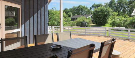 An outdoor deck with a wooden table and chairs, a white fence, and a view of a green landscape with trees and a house.