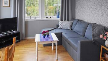 A modern living room with a grey sofa, a white coffee table with flowers, a TV on a stand, and a window with curtains.