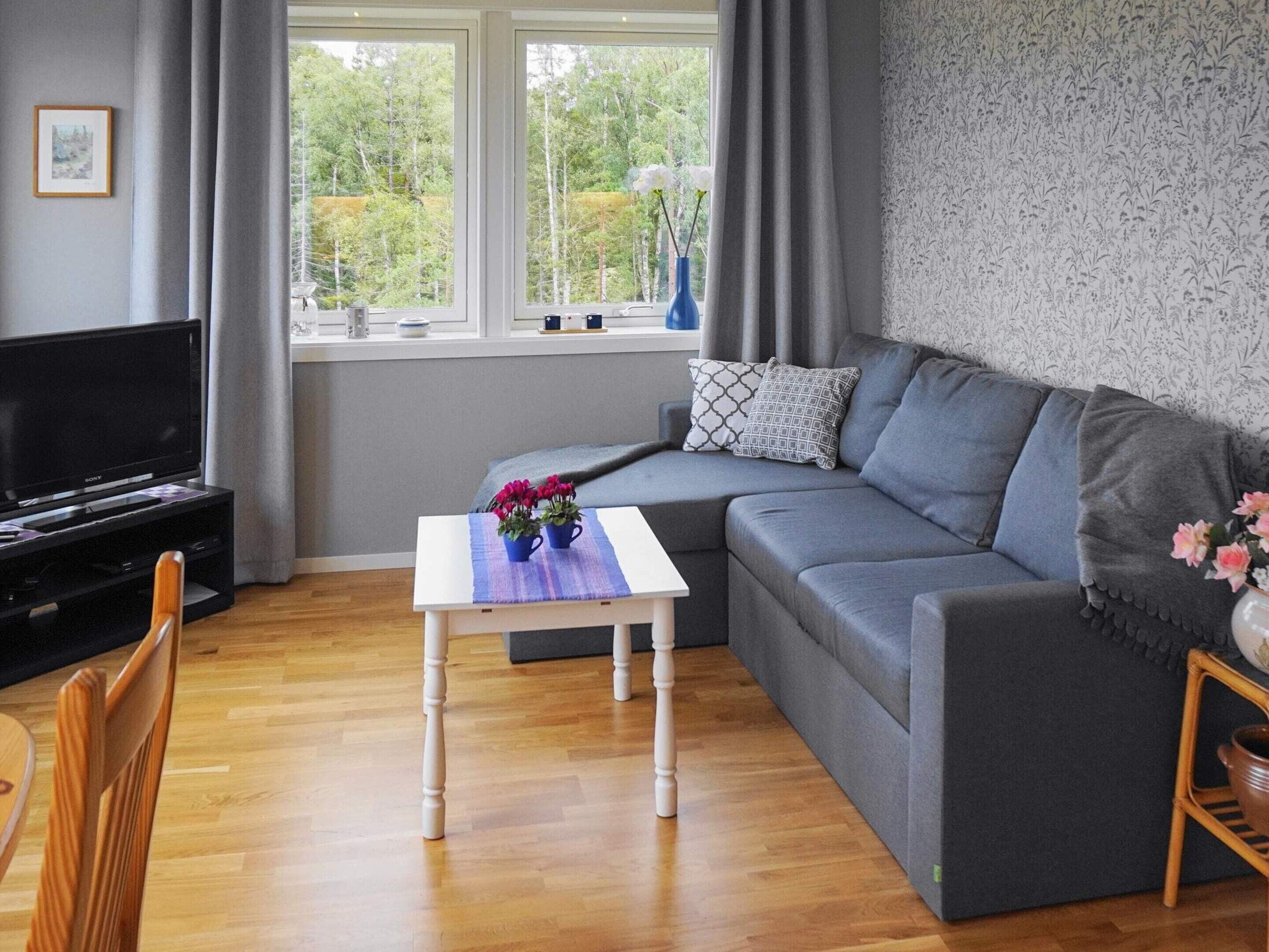 A modern living room with a grey sofa, a white coffee table with flowers, a TV on a stand, and a window with curtains.