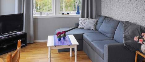 A modern living room with a grey sofa, a white coffee table with flowers, a TV on a stand, and a window with curtains.