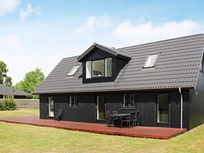 Une maison moderne avec une façade sombre, un toit en pente et une terrasse aménagée avec du mobilier d’extérieur.
