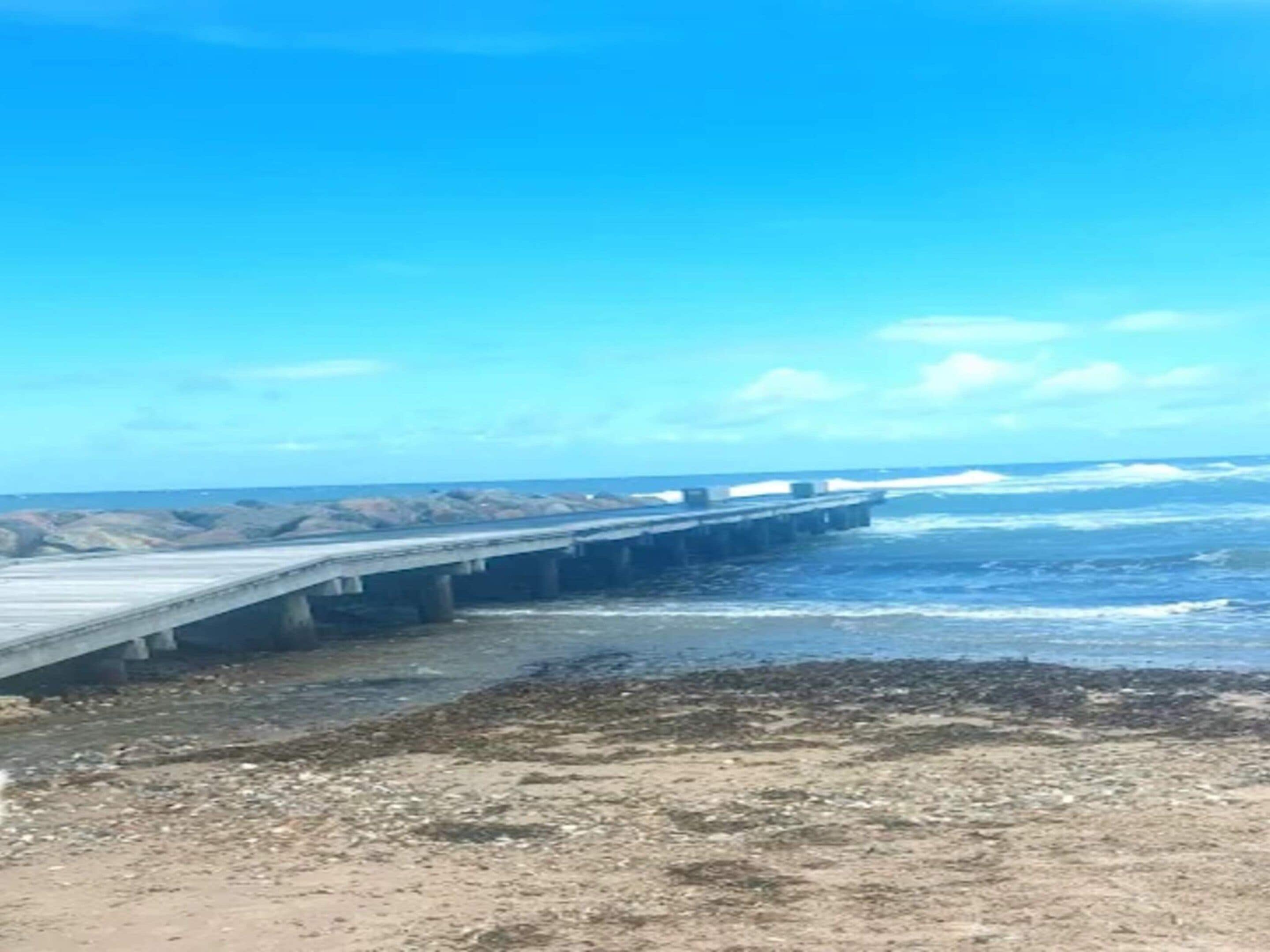 Paisaje costero con un muelle de hormigón que se adentra en el mar, terreno rocoso y cielo despejado.