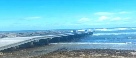 Paisaje costero con un muelle de hormigón que se adentra en el mar, terreno rocoso y cielo despejado.