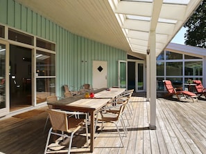 En overdækket terrasse med et bord og stole, store vinduer og en dør, der fører til et poolområde.