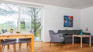 Ein modernes Wohnzimmer mit Holzdielen im Fischgrätmuster, einem Esstisch mit Stühlen, einem grauen Sofa, einem kleinen Holztisch und einem farbenfrohen abstrakten Gemälde an der Wand.