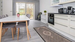 Eine moderne Küche mit weißen Schränken, ein Essbereich mit weißem Tisch und Stühlen sowie ein Blick ins Freie durch ein großes Fenster.