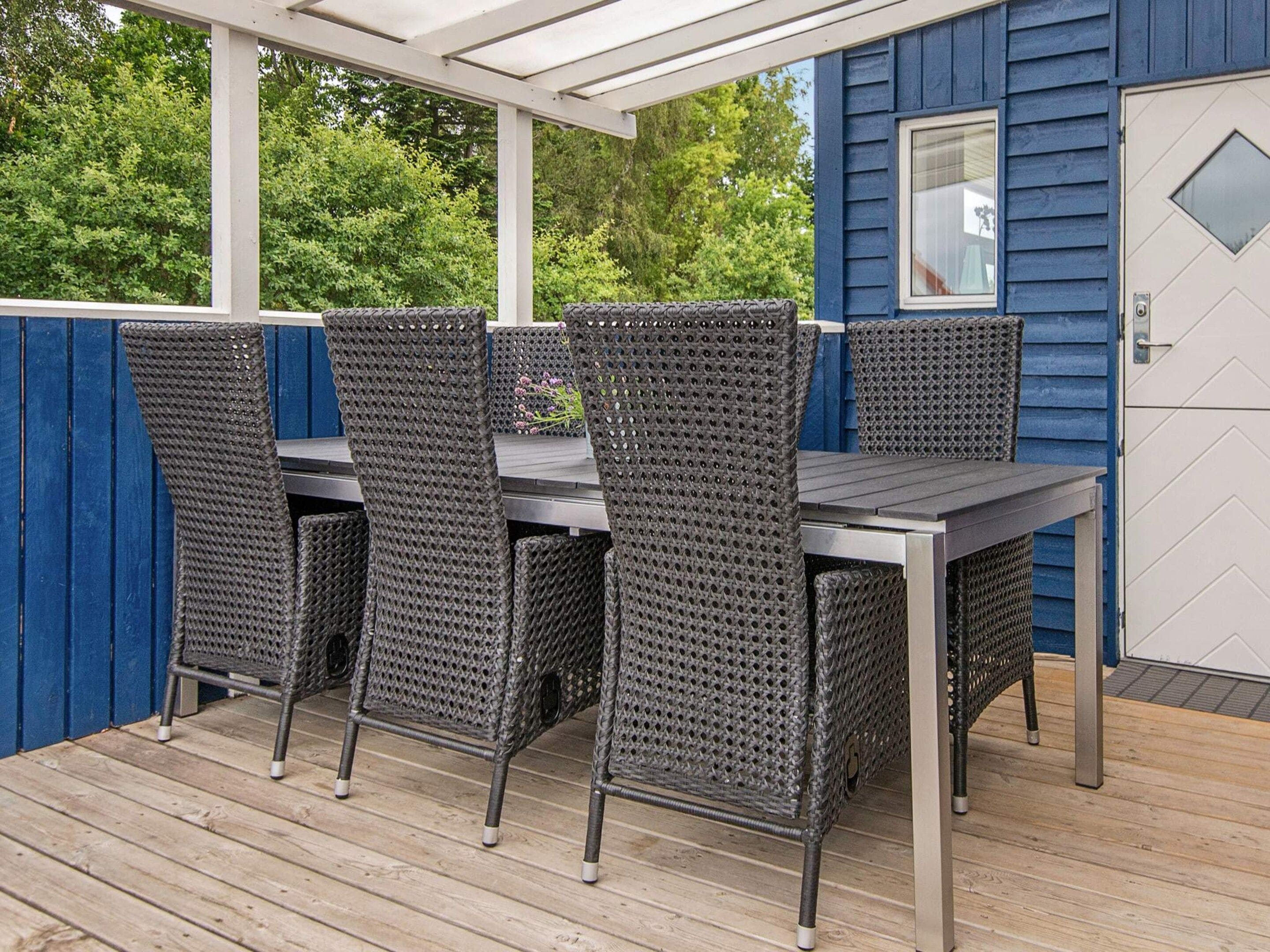 An outdoor patio with a table and four chairs, a wooden deck, and a blue building with a white door.