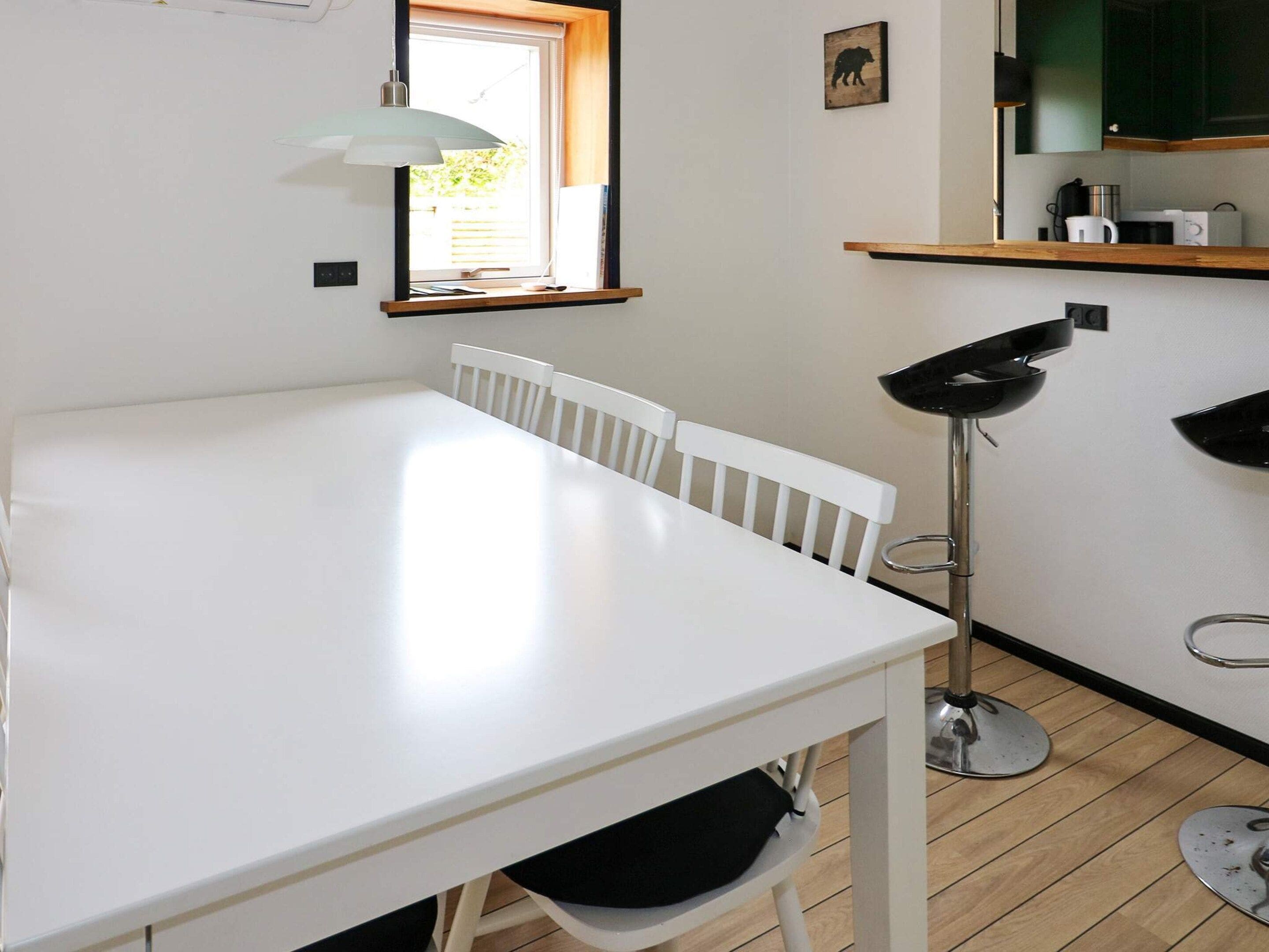 Una mesa de comedor blanca con sillas, una lámpara de pie, una ventana con persianas, y una zona de cocina con armarios y electrodomésticos.