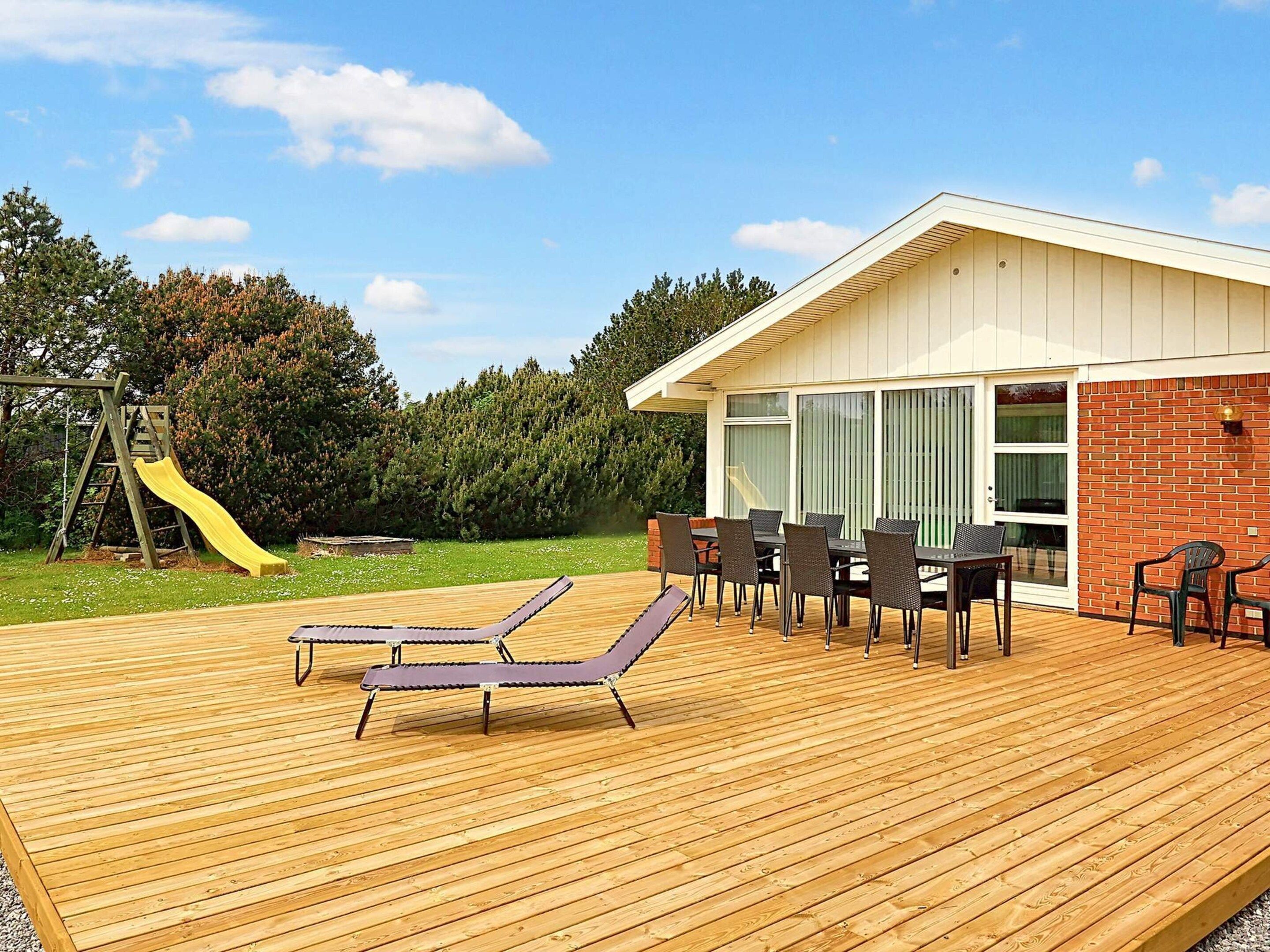 A wooden deck with lounge chairs, a dining table, and a brick building with large windows.