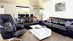 A modern living room with a black leather sofa, a white coffee table, and a large window with blinds. - 8 Person Holiday Home in Vaeggerlose (Vaeggerlose)