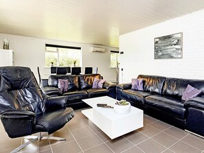 A modern living room with a black leather sofa, a white coffee table, and a large window with blinds. - 8 Person Holiday Home in Vaeggerlose (Vaeggerlose)
