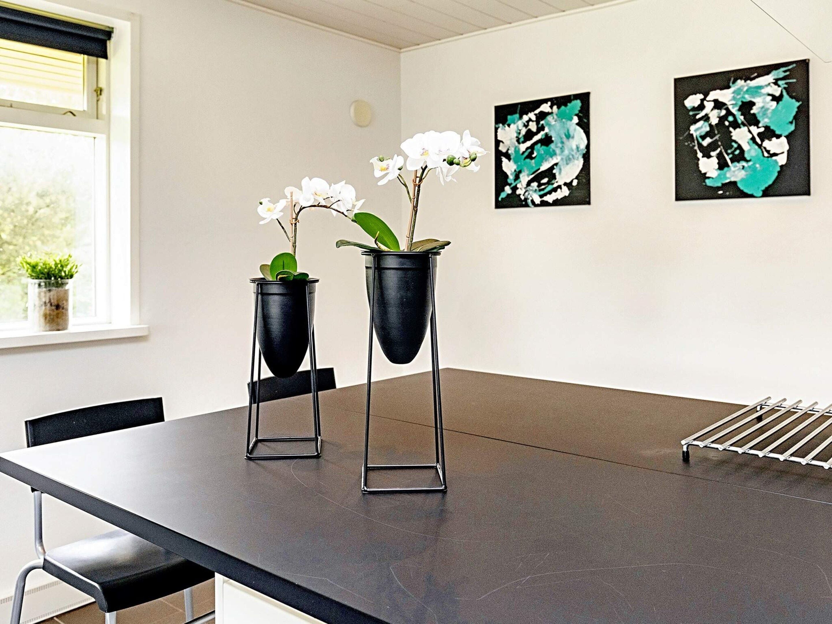 Et moderne spiseområde med et sort bord, to sorte plantekasser med hvide blomster og abstrakt kunst på væggen.