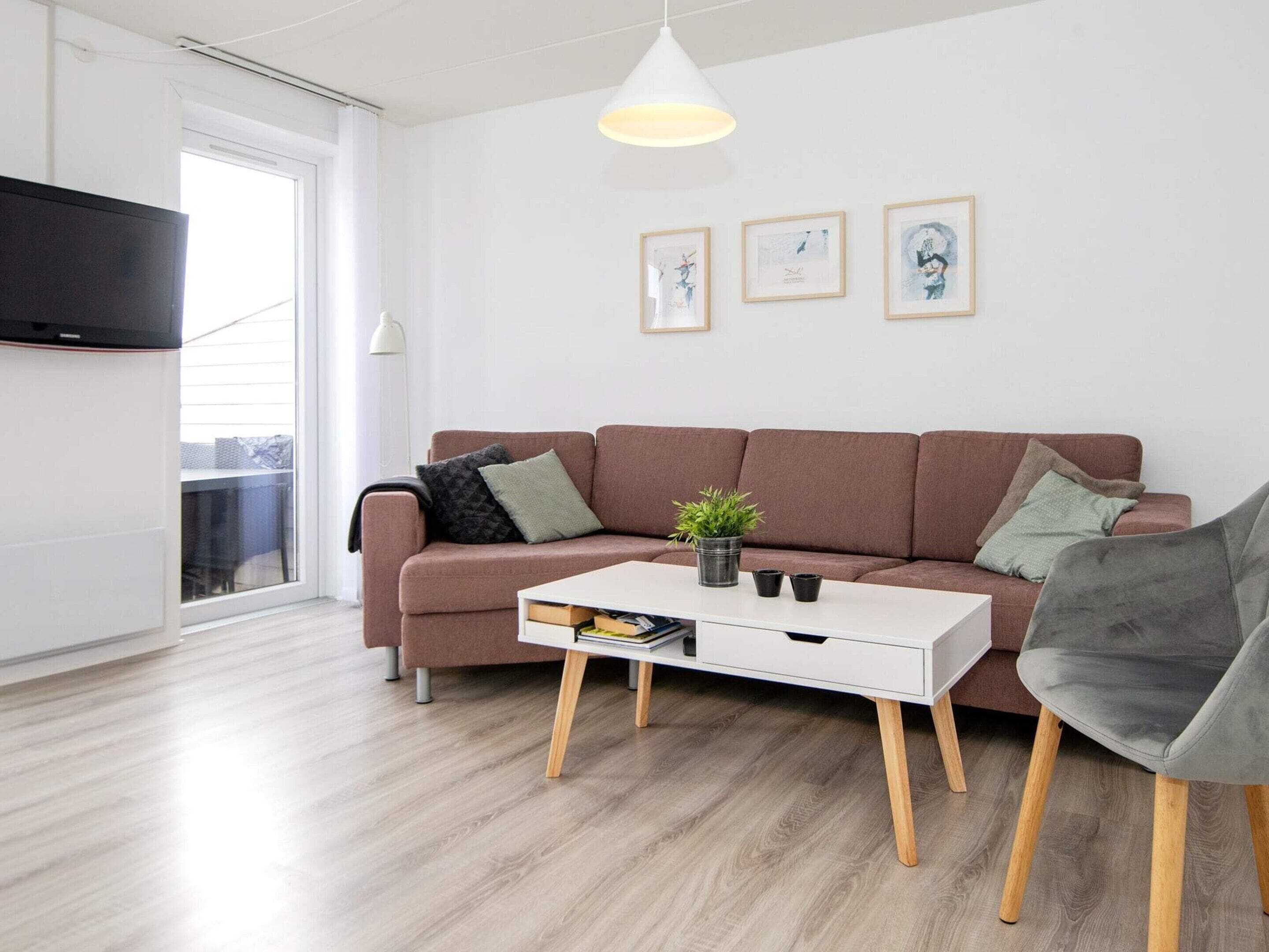 A modern living room with a sofa, a coffee table, and a grey armchair. There are framed artworks on the wall and a potted plant on the coffee table.