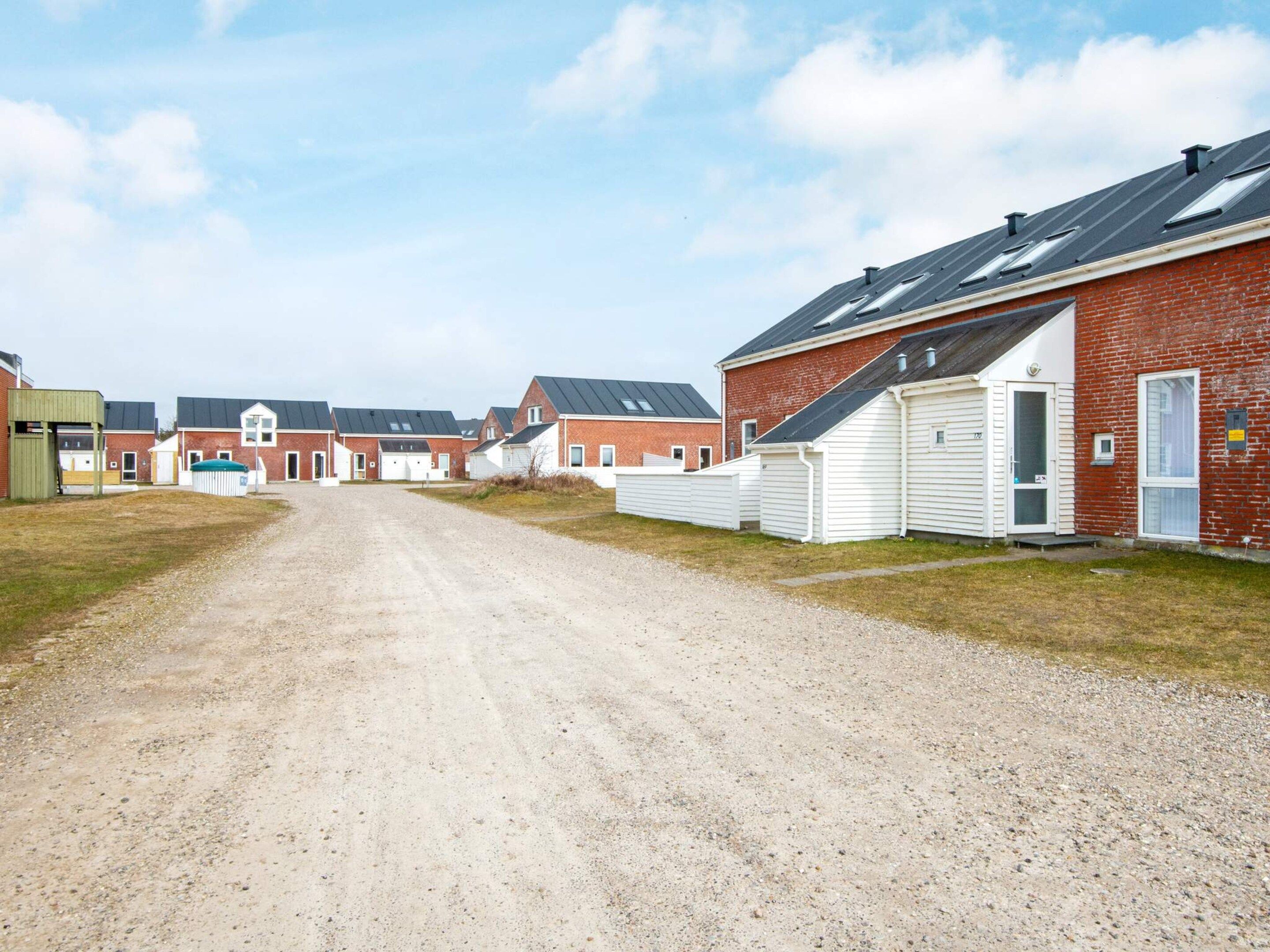 A gravel driveway leading to a row of red brick houses with white siding and a covered carport.