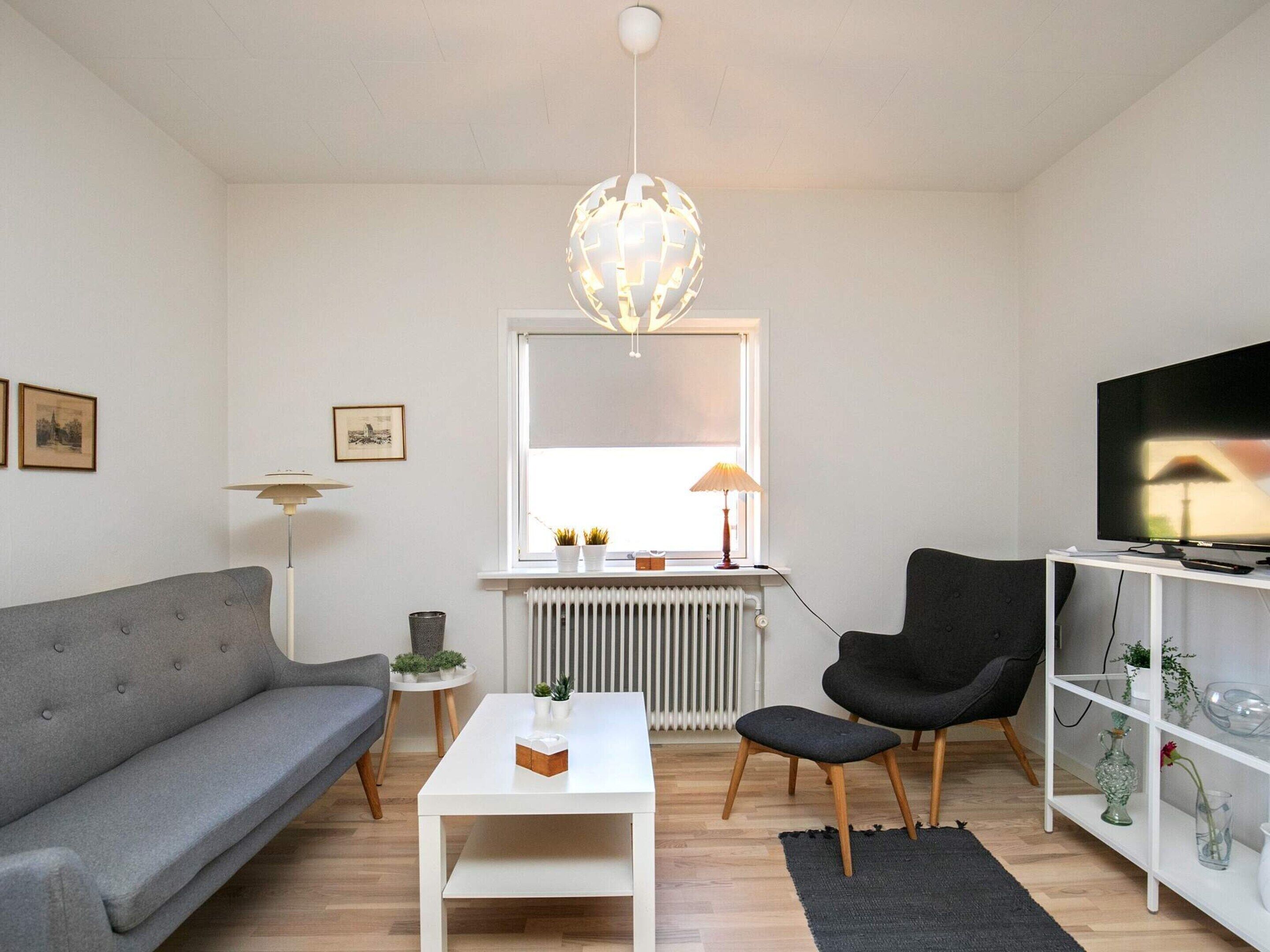 Una sala de estar moderna con un sofá gris, una mesa de centro blanca, un sillón negro, un televisor sobre un soporte blanco y una lámpara esférica original.