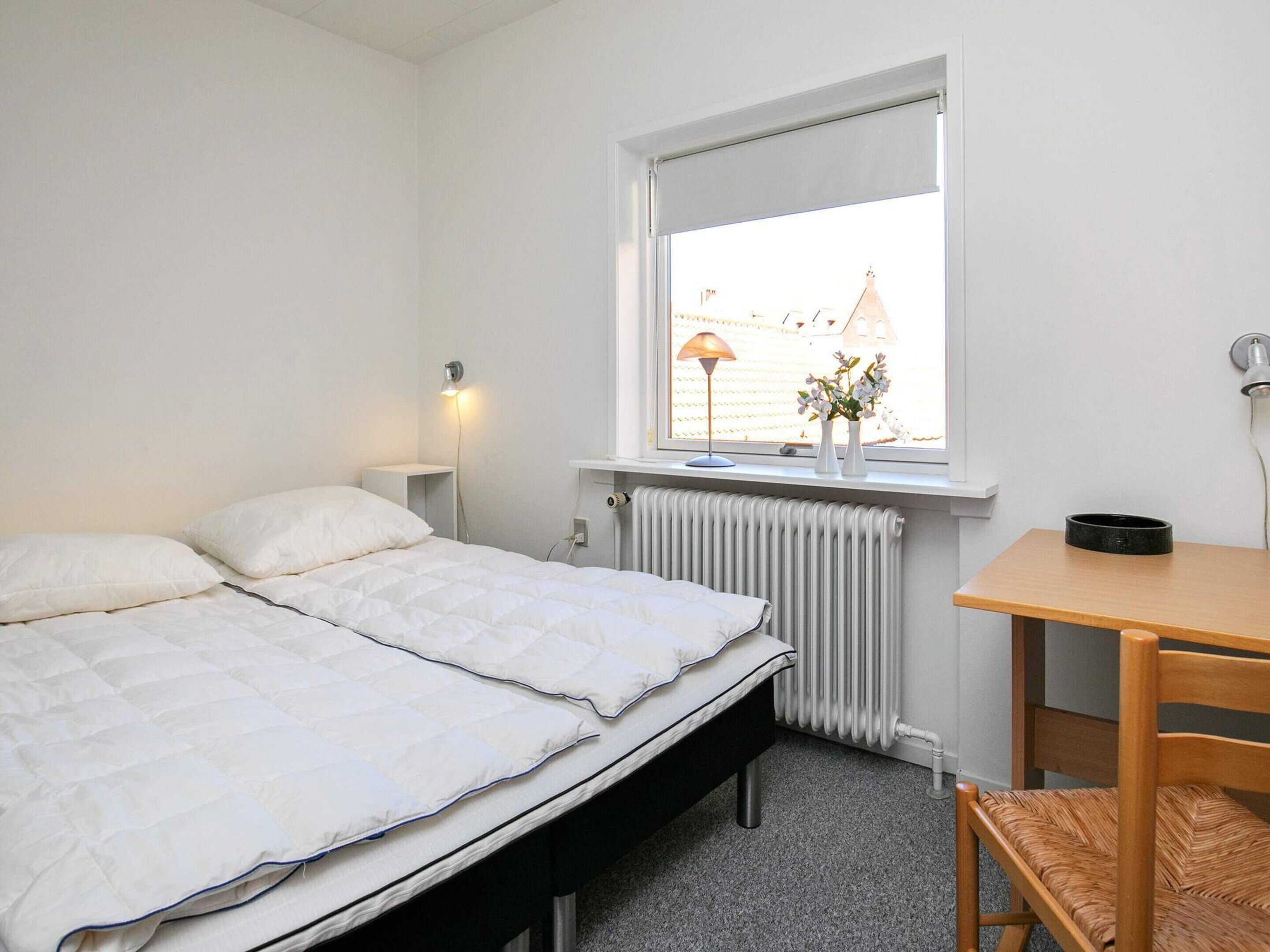 Une chambre à coucher comprenant un lit, une table de chevet, une lampe, un radiateur, une fenêtre et une vue sur les bâtiments environnants.