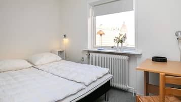 Une chambre à coucher comprenant un lit, une table de chevet, une lampe, un radiateur, une fenêtre et une vue sur les bâtiments environnants.