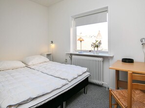 A bedroom with a bed, bedside table, lamp, radiator, window, and a view of buildings outside. - 4 Person Holiday Home in Skagen (Skagen)