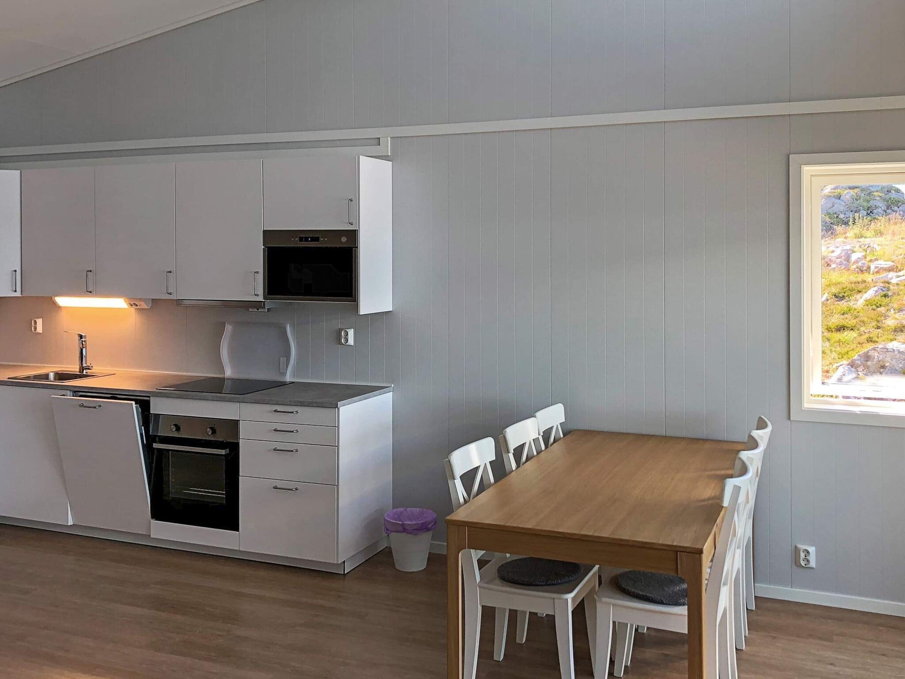 Een compacte keuken met witte kastjes, een ingebouwde oven en een eethoek met een houten tafel en witte stoelen.