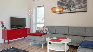 A modern living room with a grey sofa, a red cabinet, a flat-screen TV, a round white table with red accents, and a hanging lamp. - 6 Person Holiday Home in Nexo (Nexø)