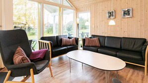 Ett modernt vardagsrum med en svart lädersoffa, ett träbord och en grå fåtölj. Stora fönster ger utsikt över omgivningen.