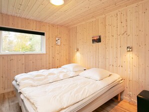 A room with a wooden paneled wall, a bed with white bedding, a window with a view of trees, and a wall-mounted light fixture. - Holiday Home in Hals (Hals)