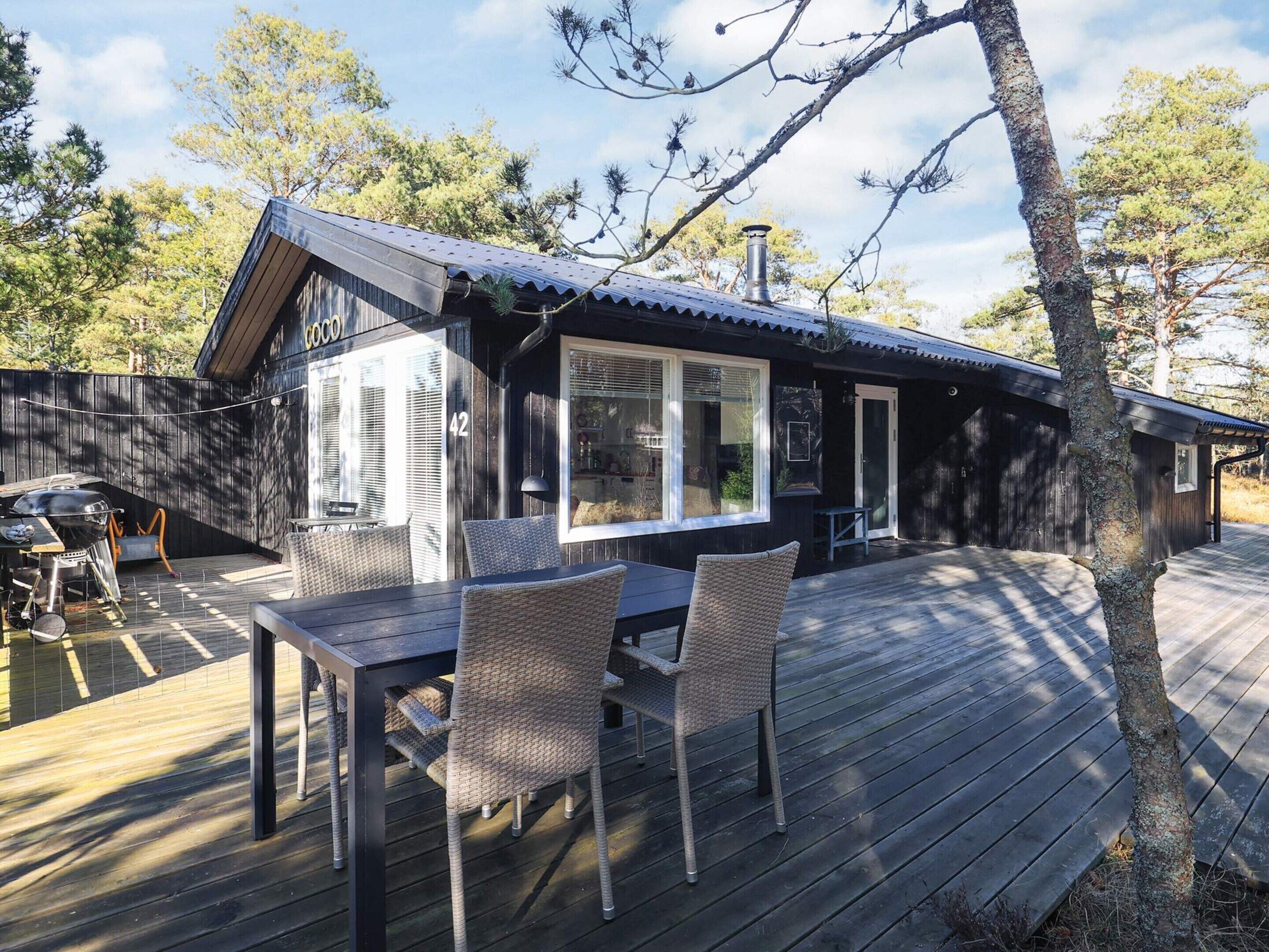 En træterrasse med to fletstole og et bord, en grill og en bygning med mørk facade og metal tag.